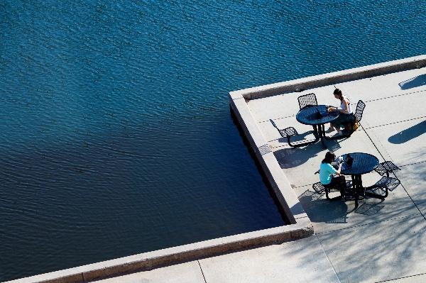  As seen from above, students sit at tables enjoying lunch along a blue pond. 