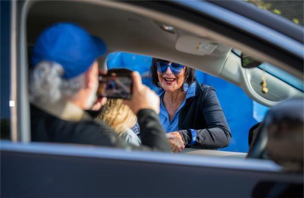 A person wearing blue glasses with heart-shaped lens reaches into a car to shake the hand of a passenger while the driver uses a phone to take a photo of the person greeting them.