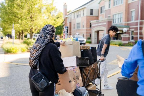 A person pulls on a cart full of items and looks back at another person carrying items.