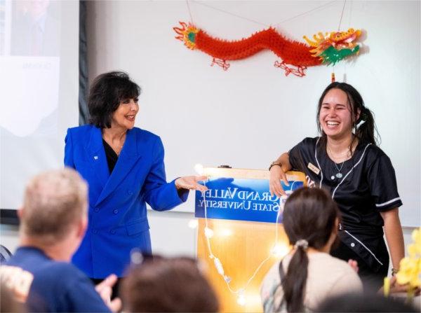 One person laughs while another gestures toward them with their hand. A podium with the words, "Grand Valley State University" is behind them.