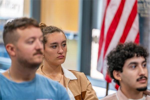 Three people look toward a speaker. An American flag is in the background.