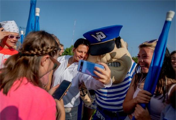 People, some of whom are holding blue flags, surround Louie the Laker. One person is taking a selfie with Louie.