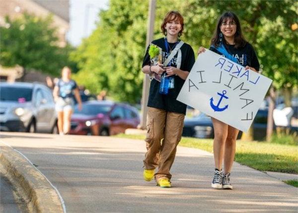 Two people smile while walking. One of them holds a sign that says "Laker Familia" with a drawing of a blue anchor.