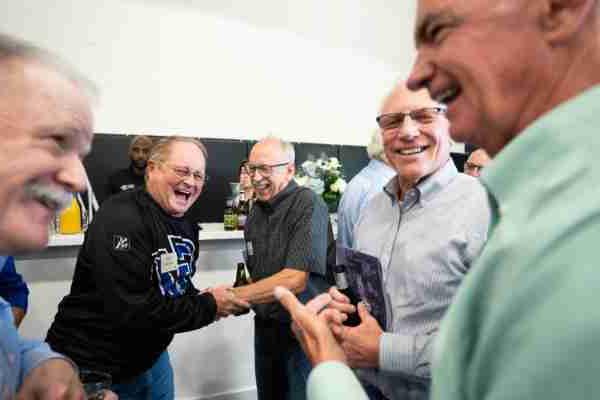 A group of older gentlemen laugh and greet each other during an event.  