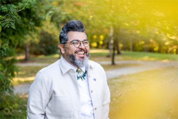   A portrait of a smiling person with glasses wearing a white jacket photographed in a colorful outdoor space. 