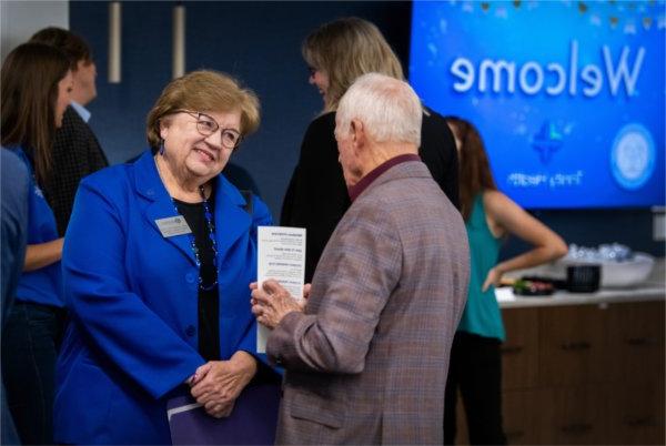 Linda Lewandowski, in blue blazer, smiling and talking with a man with gray hair, others in background at a reception