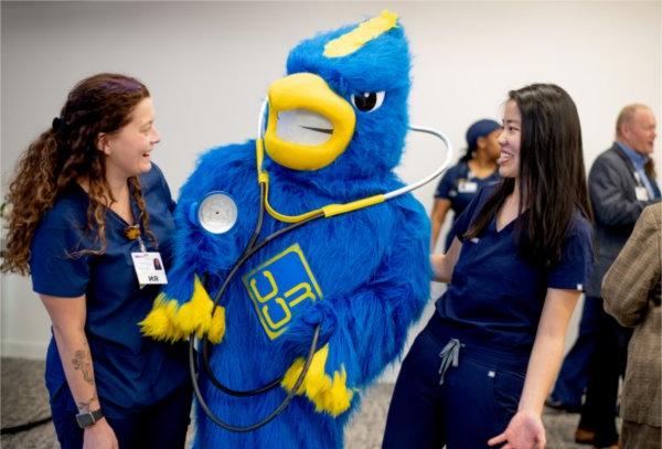 two nursing students in dark blue scrubs laugh with a jaybird mascot in center with a stethoscope around its neck