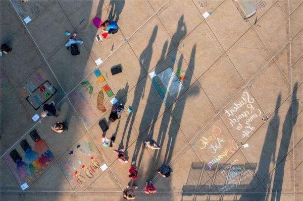  As seen from a drone, shadows of people are cast over colorful chalk art drawings on a sidewalk. 