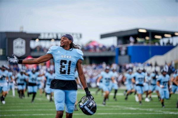  A football player holding his helmet in his hand gestures as he walks on the turf. 