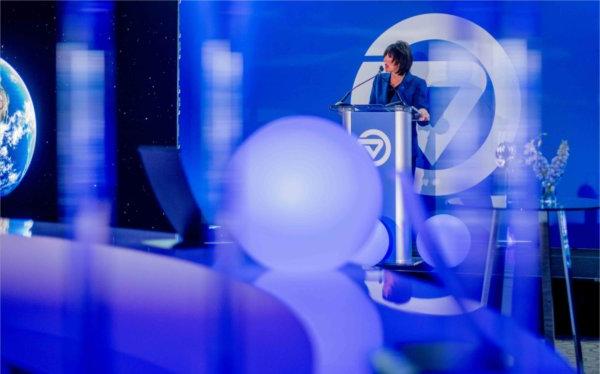  Among blue lights, a college president speaks at a lectern. 