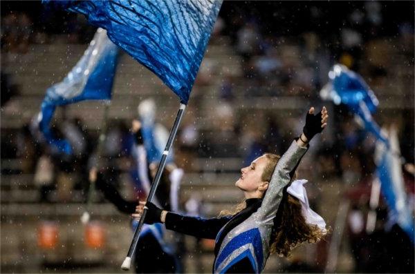 A color guard member tosses their hand in the air while twirling a blue flag in the rain.  