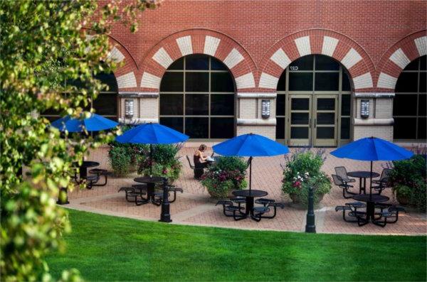  A college student works at her laptop under a blue umbrella of a courtyard on a college campus. 