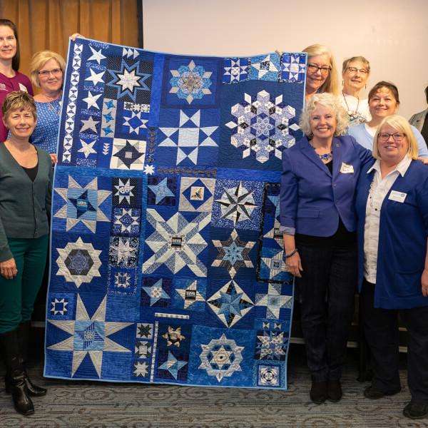 women standing in two rows surrounding a quilt