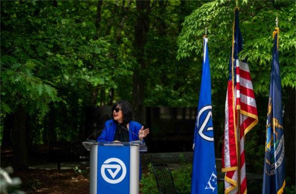 A person standing at a podium with a GVSU logo gestures with one hand while looking off to the side and speaking. Three flags are to the person's right.