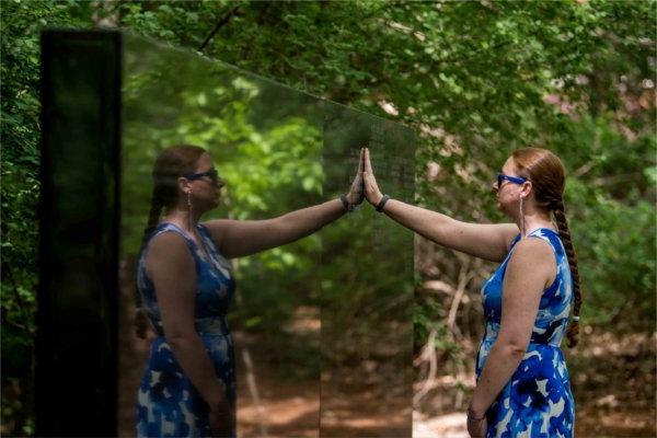 A person holds their hand to a stone wall. Their reflection is seen on the other side.