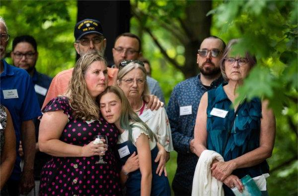 A person wraps their arm around a child, whose eyes are closed, as others look on somberly.