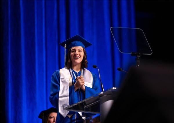  A student speaker wearing blue cap and gown speaks at the lectern. 