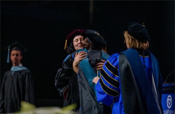 A graduate hugs a person on stage after being hooded.  