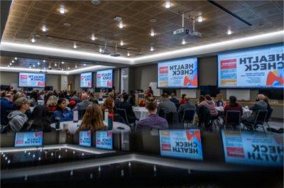 room of people seated at round tables, screens with Health Check around the room and reflected at bottom