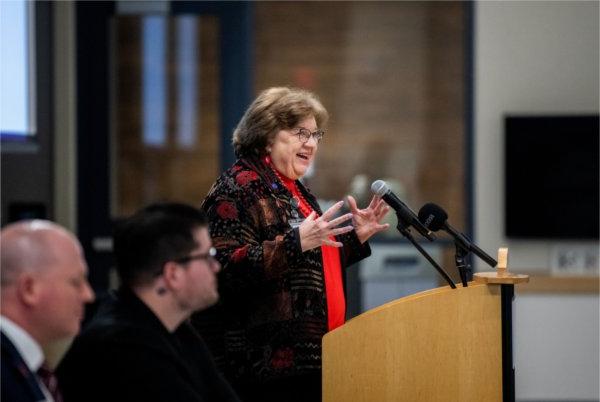 Linda Lewandowski speaking from the podium, two people seated to her left