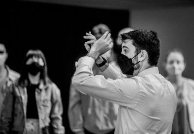 A conductor, wearing a mask, uses hand gestures while leading a choir performance.