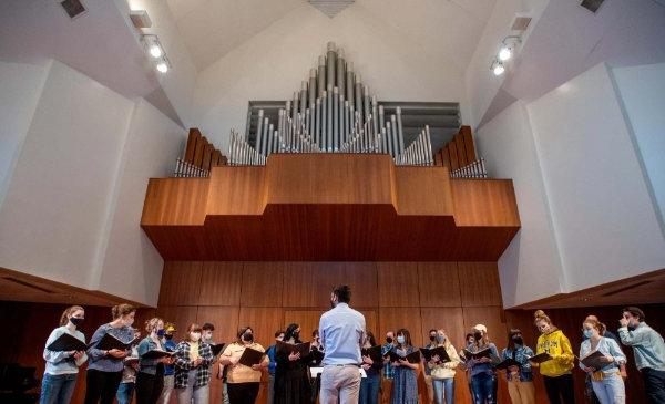 A choir performs with a conductor standing before the members, all of whom are wearing masks and holding notebooks.