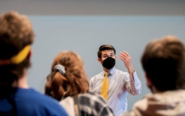 A conductor, wearing a mask, uses a hand gesture to lead choir members, some of whom are seen from behind.