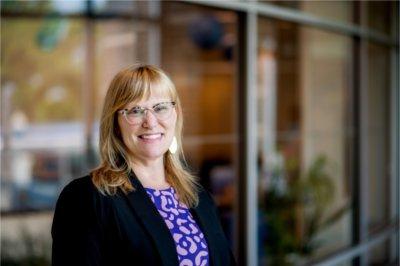 A portrait of Holly Jasinski, a woman with blonde hair and glasses. She smiles and poses in front of the Center for Women and Gender Equity.