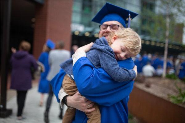  A graduate wearing cap and gown holds his toddler son before Commencement.