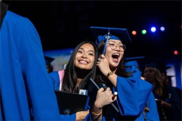  Two graduates smile and hold their graduation tassel.