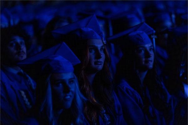  A blue light from a video casts light on the faces of graduates wearing blue caps and gowns. 