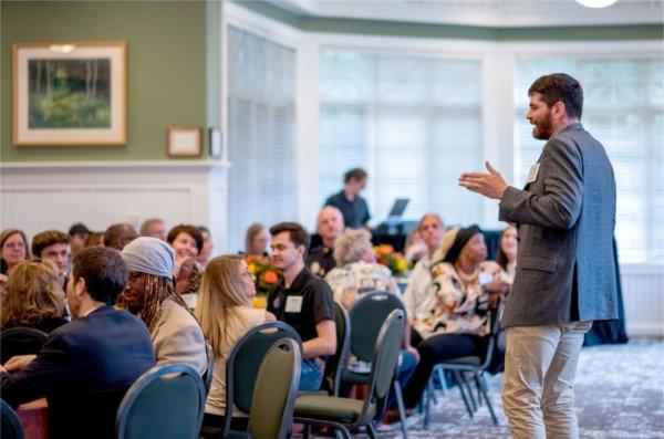 man stands to address audience seated at round tables in the Alumni House