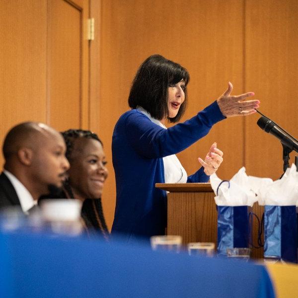 President Mantella stands behind podium, gesturing with hands to audience. Two people seated at table to her left.