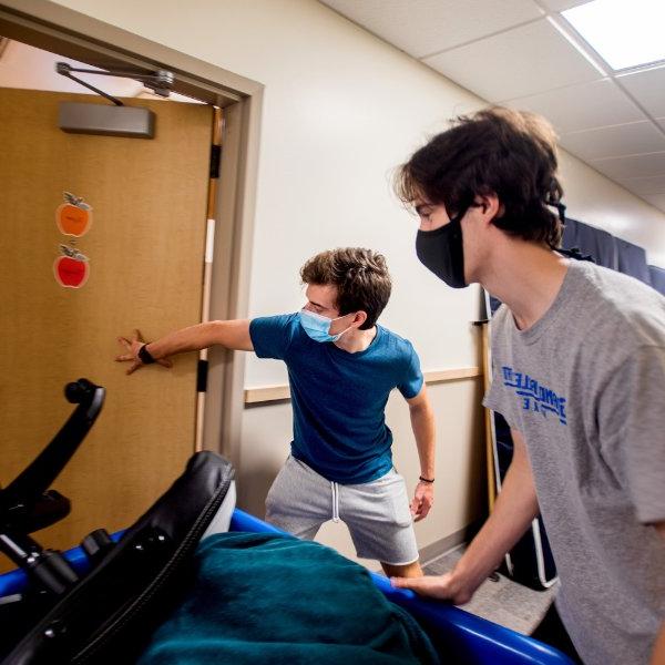 two roommates, wearing face masks, push a cart of belongings into their living center room