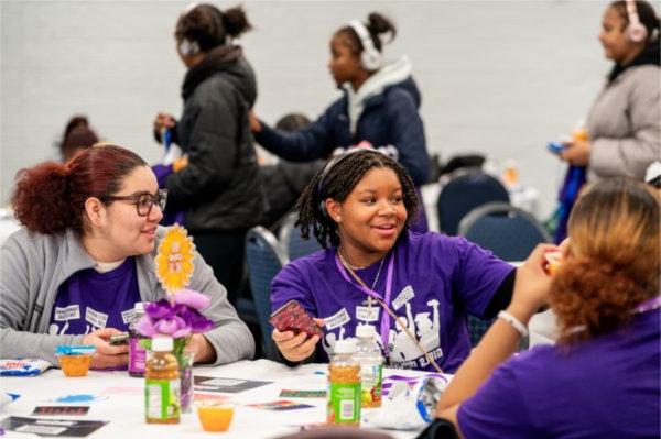 Two girls in purple shirts chat while sitting at a table.