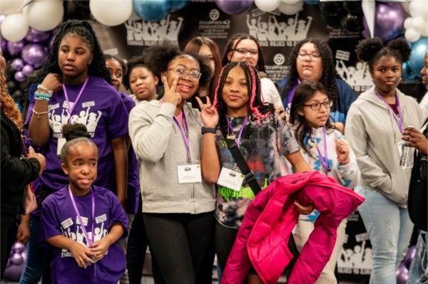 A group of girls pose for a photo in front of a Girls of Color background. The girl in the middle has pink braids and a Goosebumps shirt.