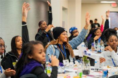 Sitting at a table, girls raise their hands and wait to be called on.